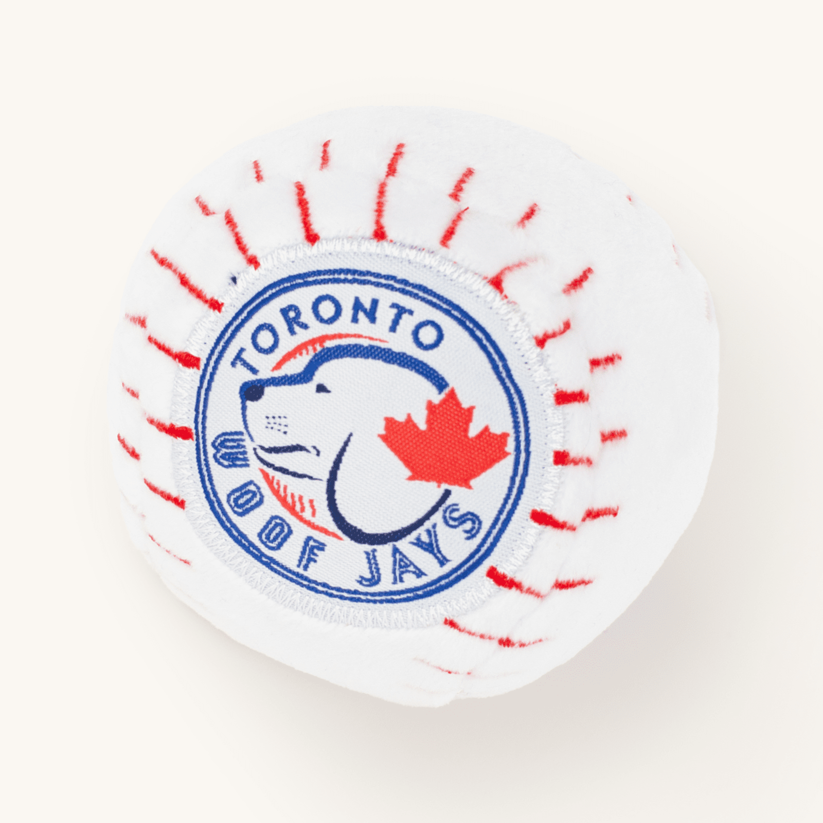 White plush ball-shaped dog toy with a Toronto sports-team patch featuring a cartoon dog and red maple leaf, on a plain cream background