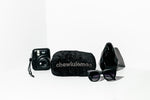 Black plush Chewlulemon belt-bag dog toy styled beside a camera, sunglasses and a dark vase on a black backdrop