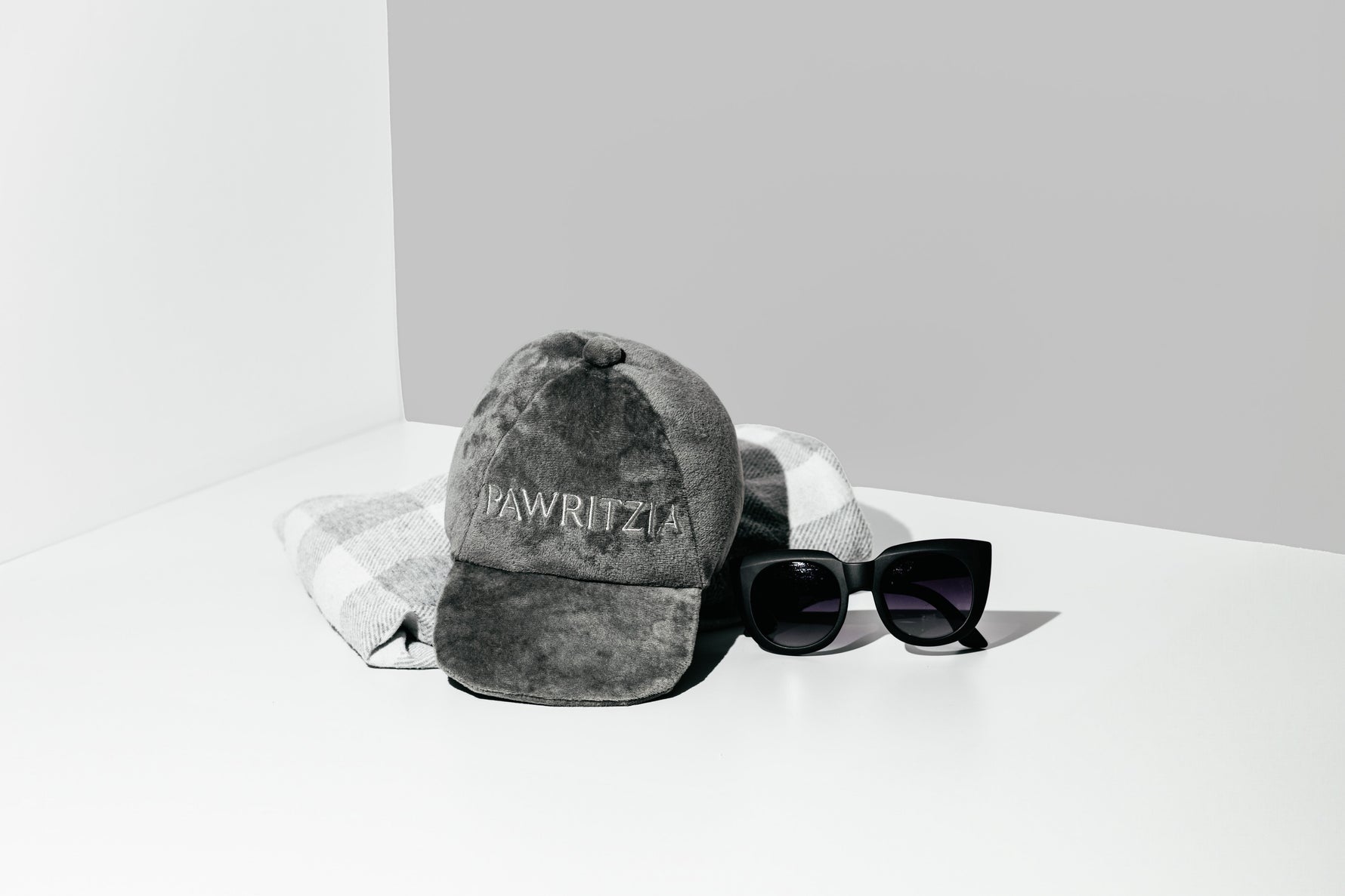 Grey plush Pawritzia cap dog toy styled with a plaid cloth and sunglasses on a grey studio backdrop